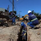 Cinco trabalhadores com uniforme e capacetes trabalham em obra com tubulações