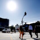 Pessoas circulam em rua com carros trafegando, no céu, é possível ver o sol