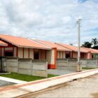 Sete casas recém-construídas, uma do lado da outra, em rua asfaltada. As casas são coloridas e tem grama na frente