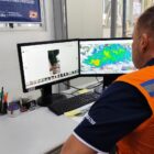 Homem com colete laranja olha para telas de computador com mapas e fotos