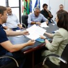 Prefeito, secretários e representantes de banco em volta de uma mesa. Prefeito entrega papel para uma mulher