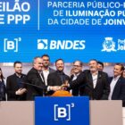 Prefeito, secretários e outras autoridades em pé, na frente de telão, alguns seguram e batem um martelo em leilão