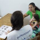 Mulher com camiseta escrito "entrevistador" conversa com mulher com criança no colo