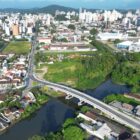 Imagem aérea da cidade, com ponte em cima de rua, área verde, prédios, casas e carros trafegando