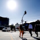 Pessoas andam em rua, carros passam, há comércios, um dia claro, com muito sol