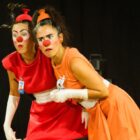 Duas atrizes, com vestidos laranja e vermelho, nariz de palhaço e luvas fazem careta em palco