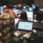 Várias pessoas viradas de costas, usando computadores, lado a lado, em auditório