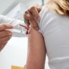 Mulher loira de costas está sentada enquanto um profissional aplica vacina em seu braço