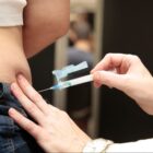 Profissional da saúde aplica injeção em mulher na parte lateral do corpo, perto da cintura