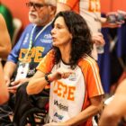 Mulher com cabelo castanha médio usa camiseta de time de basquete e fala com jogadores em cadeira de rodas