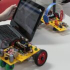 Aluna mexe em notebook ao lado de dois robôs