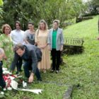 Dois homens colocam coroa de flores em túmulo de cemitério com gramado. Mulheres e um jovem observam em pé.