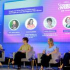 Quatro mulheres sentadas em cadeiras, em palco, seguram microfones e uma delas fala