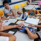 Professora ajuda alunos em tarefa, estão sentados, em roda, escrevendo em cadernos