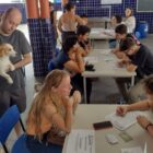 Homem em pé segura um cachorro, outras pessoas sentadas estão sendo atendidas por pessoas que escrevem em documentos
