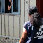 Mulher morena de costas com colete do SUS conversa com outra mulher em janela na frente de uma casa