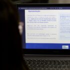 Mulher com cabelo escuro comprido de costas olha para tela de computador onde há informações sobre consulta pública