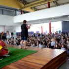 Mulher fala de palco para alunos sentados em frente. No palco, há uma menina vestida de Emília
