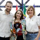 Um homem e duas mulheres (uma morena e outra loira) posam para foto. Uma delas segura troféus