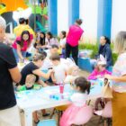 Crianças sentadas em cadeiras desenham, elas estão em praça e ao redor há adultos