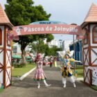 Duas mulheres atrizes com figurino sorriem e levantam os braços posando para foto em portal de Páscoa