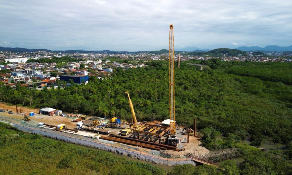 Imagem aérea de obra de ponte, há guindastes e outros equipamentos