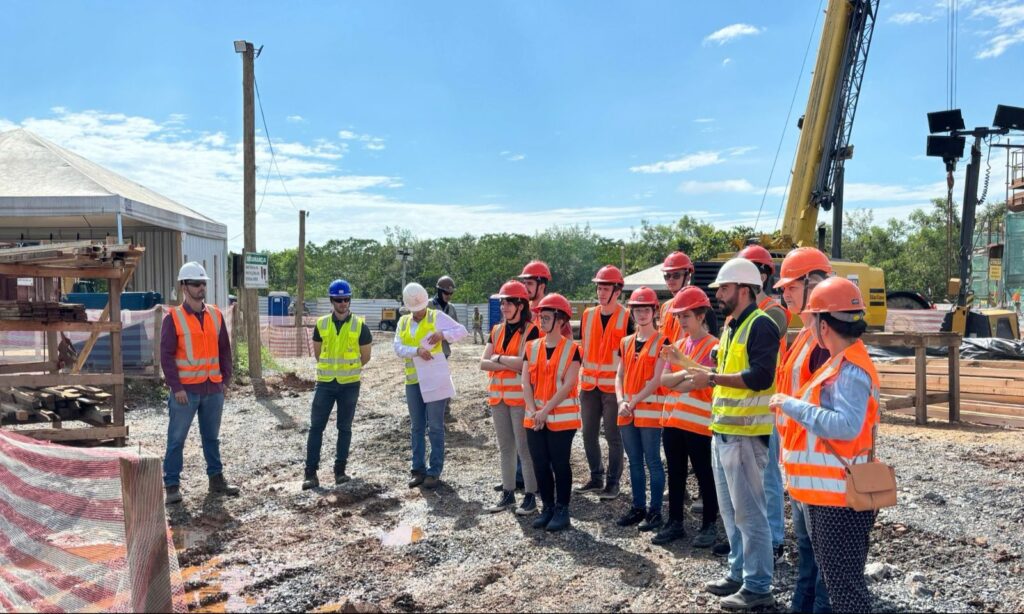 Estudantes de colete refletivo laranja e capacetes em canteiro de obras observam um engenheiro falar