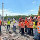 Estudantes de colete refletivo laranja e capacetes em canteiro de obras observam um engenheiro falar