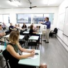 Pessoas sentadas em cadeiras na frente de carteiras olham para frente de sala onde homem em pé palestra