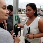 Mulher cega com bengala aciona botoeira em poste ao lado de outra mulher, um homem observa