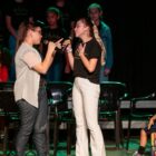 Duas meninas cantam segurando microfones em palco, uma olha para a outra