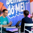Homem conversa com duas pessoas, todos estão sentados em cadeiras, em volta de uma mesa