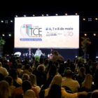 Palco de evento, com palestrantes em pé e sentados. Na frente, grande plateia sentada