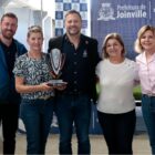 Dois homens e três mulheres posam para foto, uma delas segura um troféu