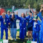 Mulheres com uniformes azuis comemoram sorrindo com braços par cima, uma segura um troféu