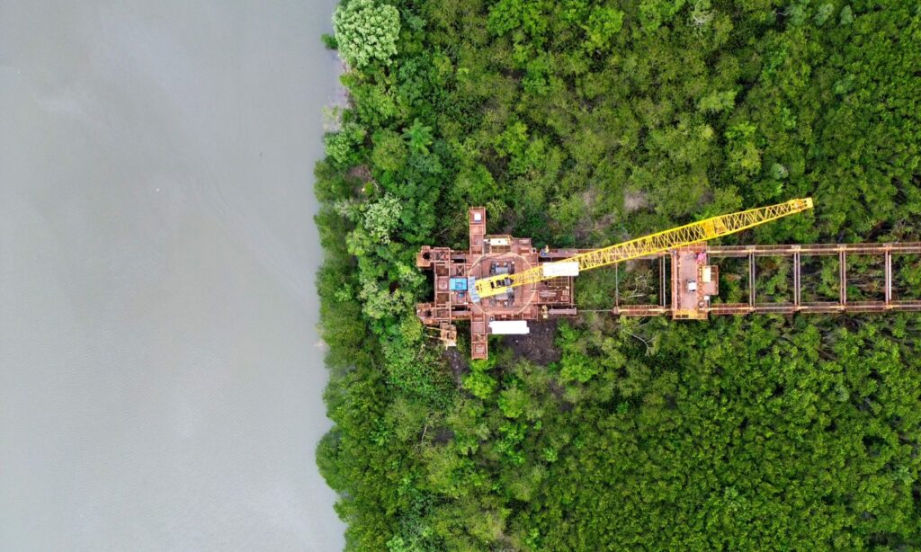 Imagem aérea de construção de ponte, com equipamentos como guindaste em margem do rio