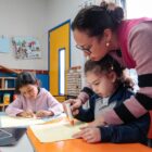 Professora segura mão de aluna na realização de tarefa em sala de aula
