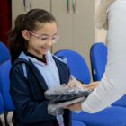 Menina com rabo de cavalo e óculos recebe kit uniforme e sorri