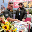 Dois homens em stand turístico mostram material a outro homem sobre atrativos de Joinville