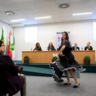 Mulher apresenta dança com roupa cigana em evento, na frente de mesa com autoridades