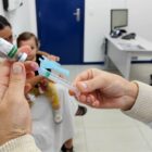 Profissional segura seringa e frasco com vacina. Mãe com filho no colo sentada na sala para receber a vacina