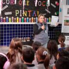 Mulher em pé segura microfone e cartaz e fala com alunos sentados