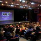 Pessoas sentadas em plateia de teatro em frente a palco com telão sobre campanha