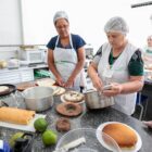 Três mulheres com avental e touca em cozinha olham e manuseiam panela, ao lado de pães e bolos