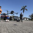 Dois homens, um com uniforme da Defesa Civil e outro da Guarda Municipal, usam drones em área aberta, perto de viaturas