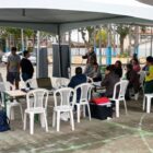 Embaixo de uma tenda pessoas sentadas em cadeiras trabalham e outras esperam para ser atendidas