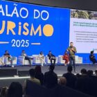 Homem palestra com microfone na mão em palco na frente de telão com plateia sentada e pessoas no palco sentadas