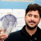Homem de cabelo castanho e barba segura um frasco fechado com mosquitos e sorri