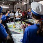 Alunos uniformizados de azul estão em pé ao lado de mesa onde robôs são avaliados