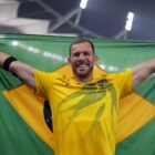 Homem com cabelo e barba castanho claro sorri, segura bandeira do Brasil de braços abertos